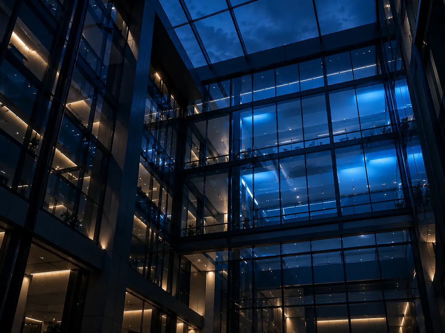 Looking up into a glass atrium at dusk, its grid of panels partly lit in blue.