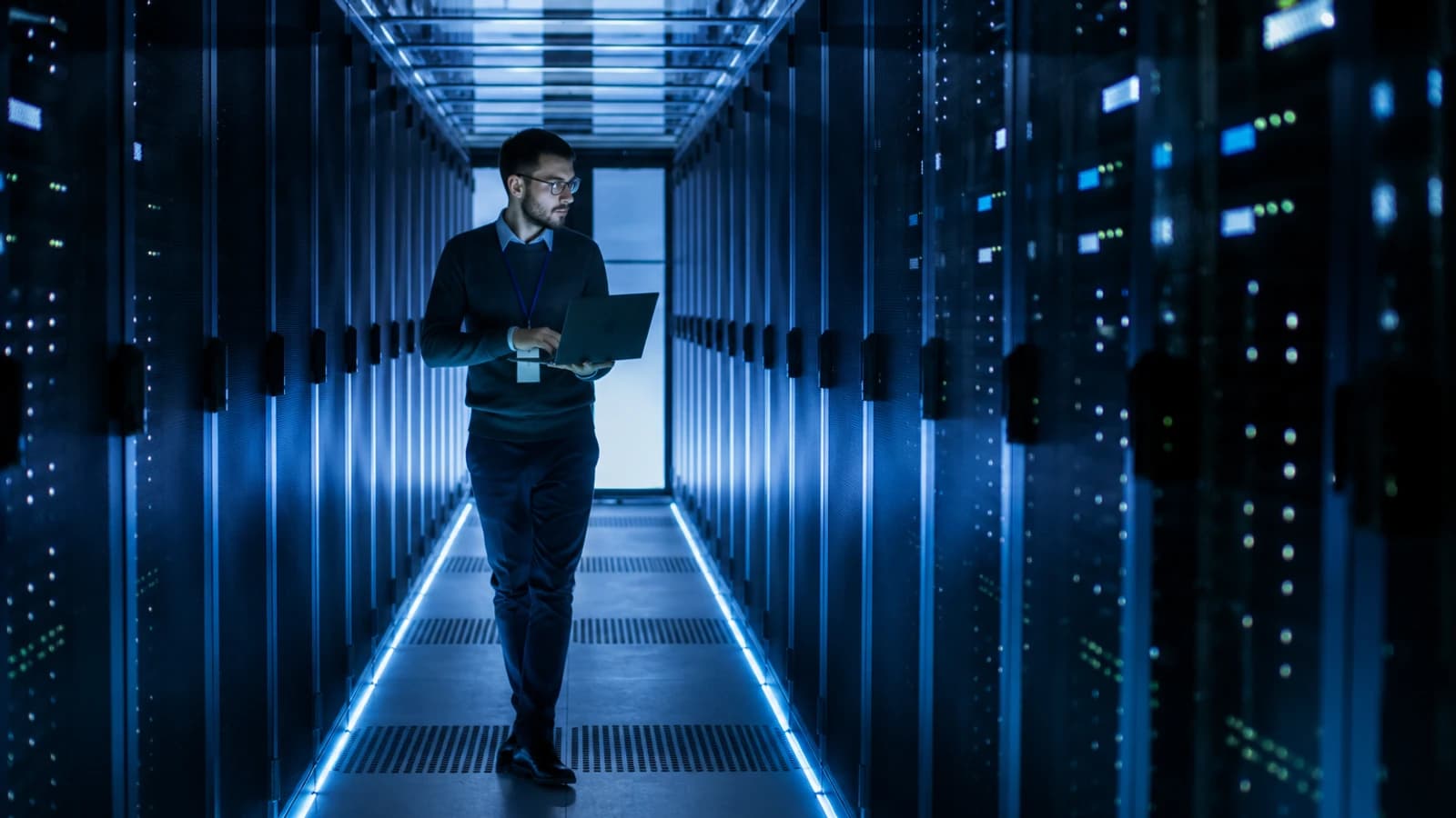 A man carrying a laptop walks down a server-room aisle lit in blue.